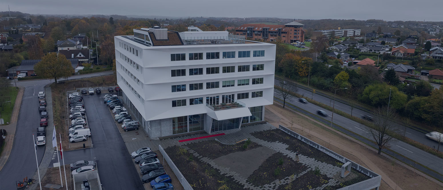 Luftfoto af en moderne hvid kontorbygning med parkerede biler og nærliggende veje på en overskyet dag.